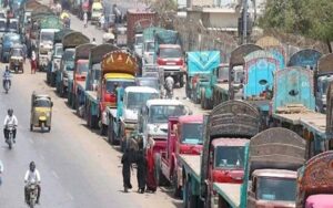 A long line of trucks parked on a busy street, with pedestrians and buildings in the background. Multiple trucks lined up on a bustling street, showcasing urban activity and traffic congestion. A busy street filled with a long queue of trucks, illustrating heavy traffic and urban logistics.